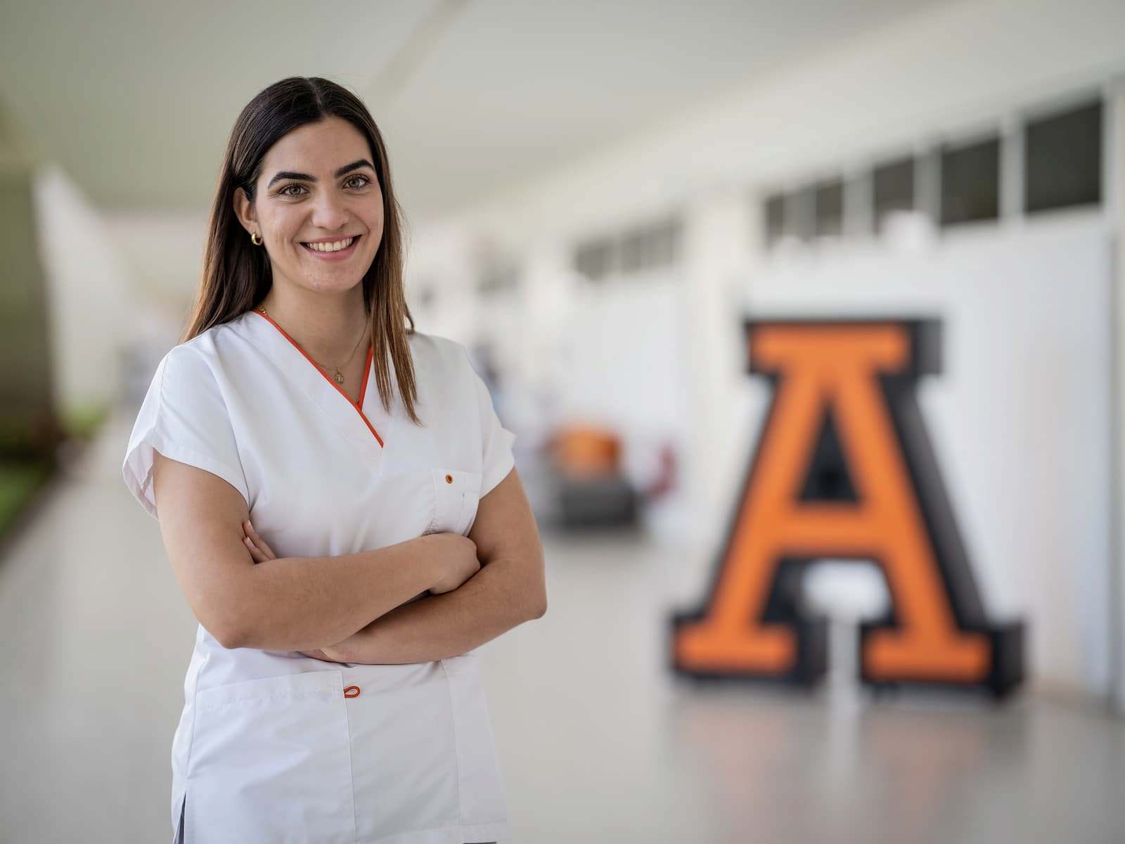 Estudiante con uniforme blanco de ciencias de la salud sonríe de brazos cruzados frente a una escultura grande de la letra 